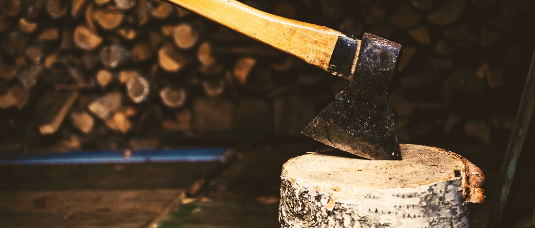 An axe on a tree trunk with stacks of logs in the background