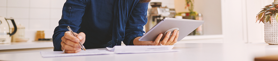 A person reading a tablet while writing on a notebook in a kitchen