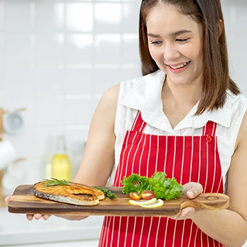 An image of a woman holding cooked fish and other vegetables on a wooden board