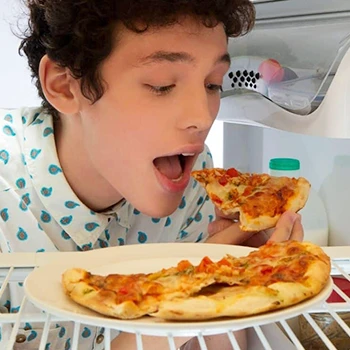 A person eating a pizza slice in front of the fridge